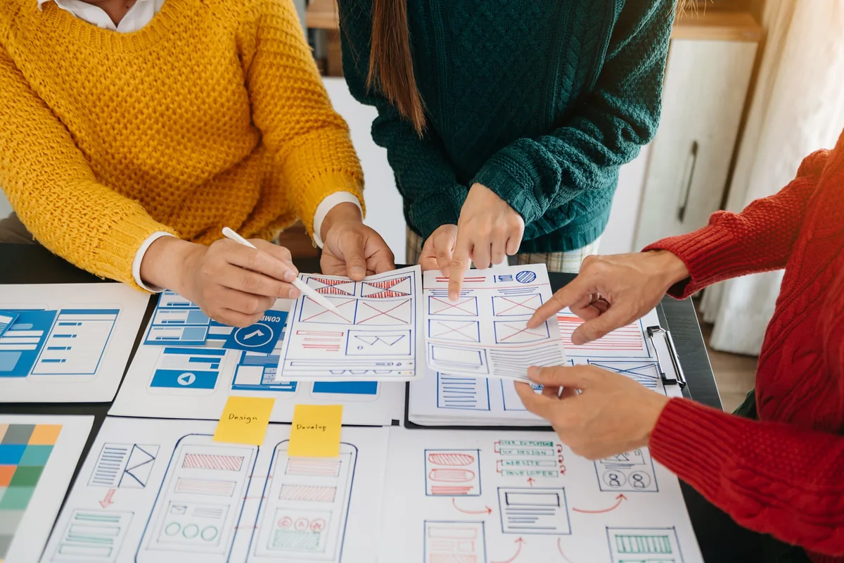 Three people point at printed app and website wireframes spread across a table