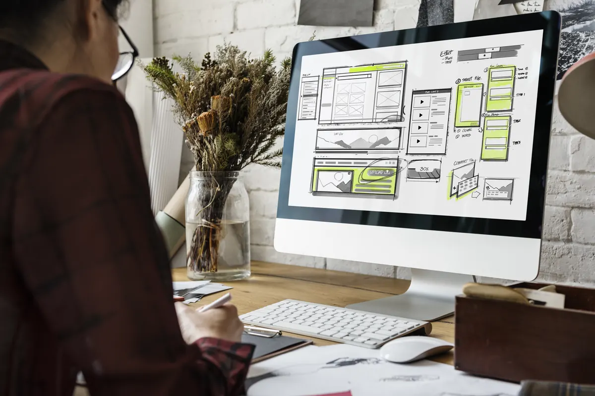 A designer at a desk reviews a website wireframe on a large monitor, beside a vase of dried flowers