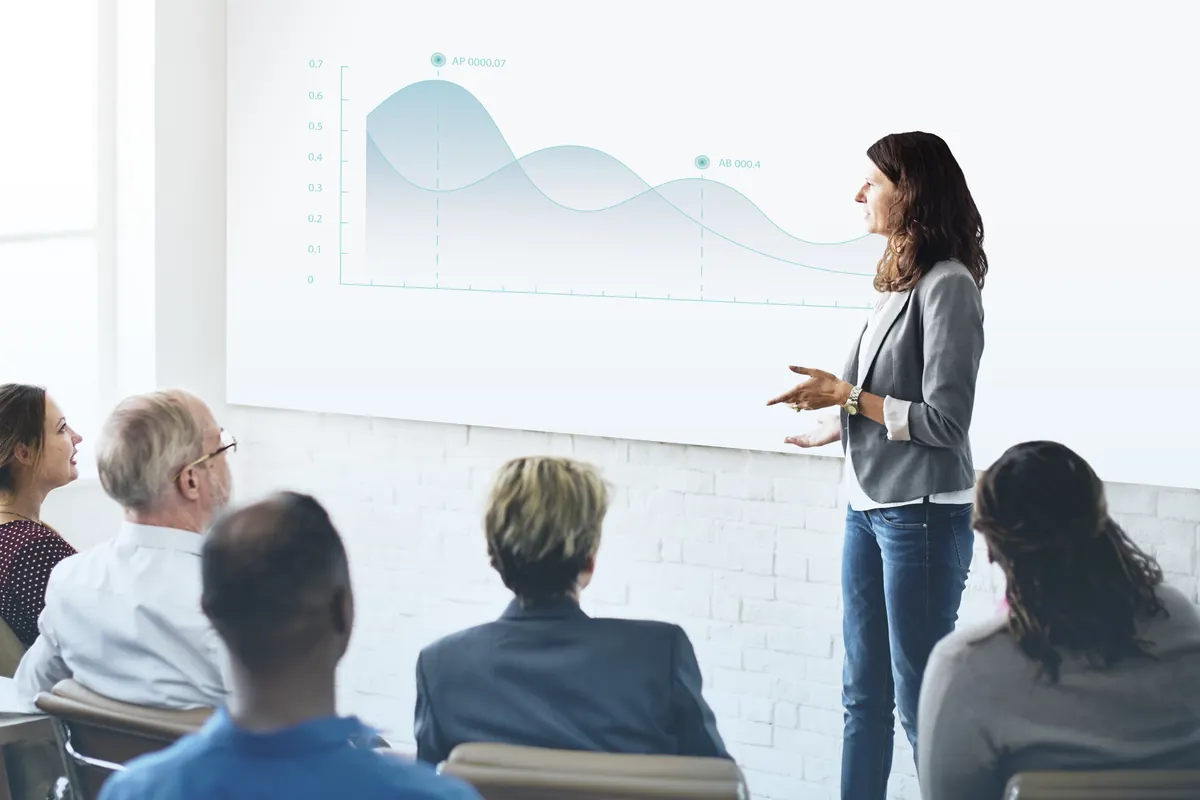 A presenter walks a seated group through a chart in a bright meeting room